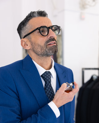 Un uomo con gli occhiali e un completo blu tiene in mano un flacone di profumo, in un negozio luminoso.