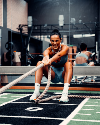 Un’atleta in squat tira una corda da battaglia, ridendo, in una palestra con il pavimento verde e nero.