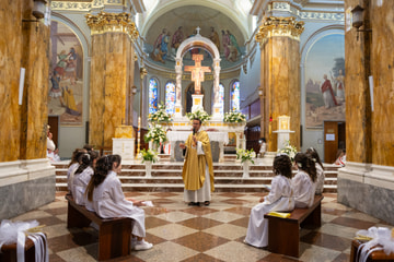 L’interno di una chiesa luminosa: l’altare con il Crocifisso e il sacerdote che parla ai bambini seduti sulle panche, in abito bianco.