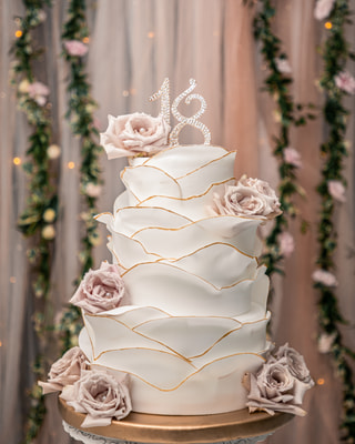Torta a piani di pasta di zucchero con il bordo dorato, rose color cipria e un «18» di strass, davanti a un tendaggio rosa con tralci d’edera e lucine.