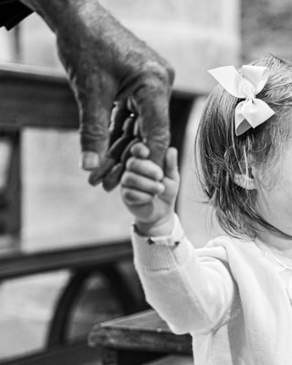 In bianco e nero, la mano segnata di un nonno stringe quella di una bambina vista da dietro, con un fiocco tra i capelli.