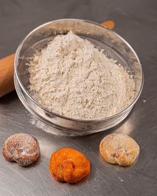 Un setaccio di farina, un mattarello e tre dolci fritti passati nello zucchero su un piano d’acciaio.