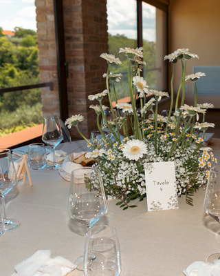 Una tavola apparecchiata con un centrotavola di margherite e gypsophila, davanti alla vetrata sulle colline.