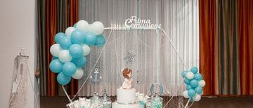 Un tavolo dolci con una torta a due piani, vasi di confetti e un arco di palloncini turchesi e bianchi, davanti a una rete da pesca e a una tenda chiara.