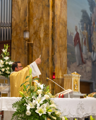 Il sacerdote in casula dorata alza l’ostia sull’altare, tra i gigli bianchi e una colonna di marmo.