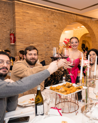 Un brindisi a una tavola rotonda: gli ospiti alzano i calici di vino rosso, con la festeggiata dietro, sotto gli archi di mattoni della sala.