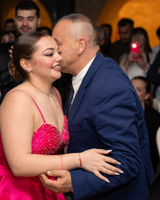 La festeggiata in abito fucsia accolta con un abbraccio da un invitato in giacca blu, tra gli altri ospiti.
