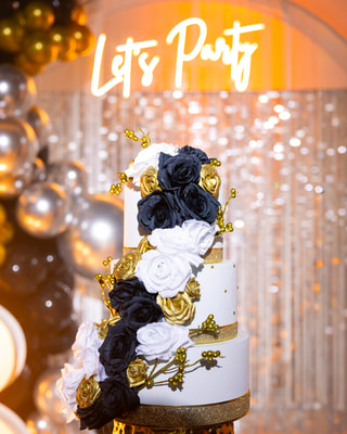 Una torta a tre piani con rose bianche, nere e dorate, davanti all’insegna luminosa «Let’s Party» e a un arco di palloncini argento e oro.