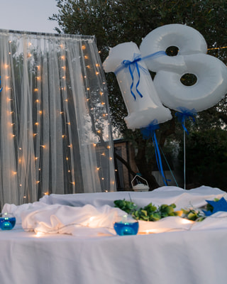 Un grande palloncino bianco a forma di 18 con nastri azzurri, una tenda di tulle con le lucine e un tavolo con le candele, in un giardino all’imbrunire.