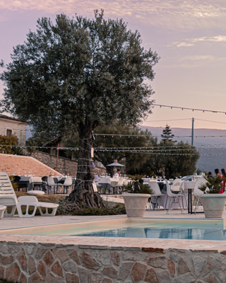 Una terrazza con la piscina al tramonto, sotto un ulivo e una fila di lucine, con le colline in fondo.