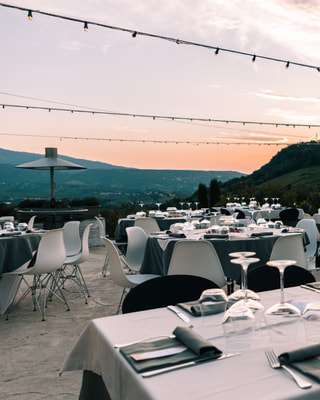 Tavoli apparecchiati su una terrazza al tramonto, sotto le lampadine, con le colline dietro.