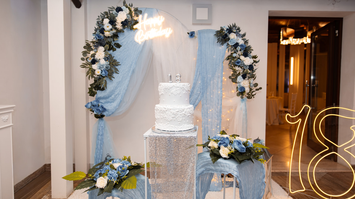 Un angolo torta in azzurro: un arco di fiori e tulle, una torta a due piani con il «18» e un «18» al neon appoggiato a terra.