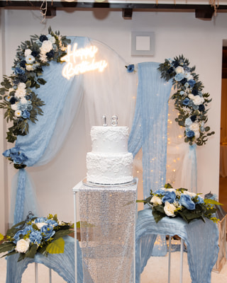 Un angolo torta in azzurro: un arco di fiori e tulle, una torta a due piani con il «18» e un’insegna luminosa.