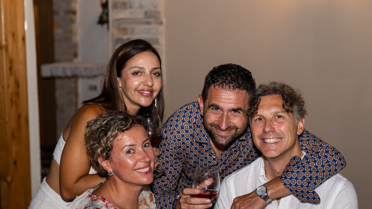 Quattro amici sorridenti stretti a una tavola, con un bicchiere di vino in mano.