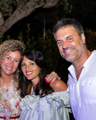 Due donne e un uomo sorridono vicini, di sera, in un giardino: una con i capelli ricci, una con i capelli scuri e lui con la camicia bianca.