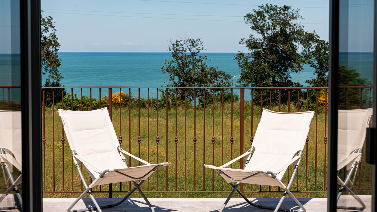 Due sdraio bianche su un balcone, viste da una porta-finestra, davanti al mare.