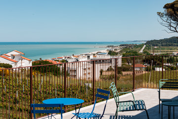 Una terrazza sulla costa con un tavolino e sedie blu e verdi, la ringhiera in ferro e il mare in fondo.