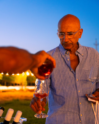 Un uomo con gli occhiali, in camicia a righe, assaggia un calice di vino rosato all’ora blu, con le lucine accese dietro e una mano che versa.