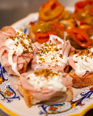 Crostini con mortadella, stracciata e granella di pistacchio su un piatto di ceramica decorata, con peperoncini sott’olio.