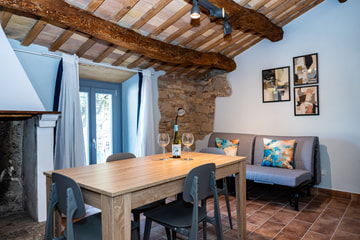 Un soggiorno con il soffitto a travi, una parete di pietra, un tavolo di legno con una bottiglia e due calici e un divano grigio con i cuscini colorati.