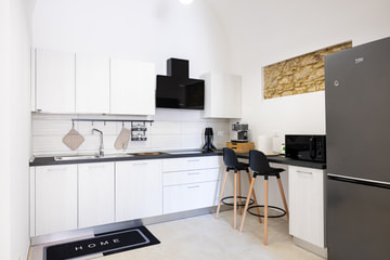 Una cucina bianca e ordinata con il piano scuro, due sgabelli neri al bancone e il frigorifero grigio.