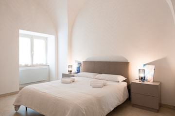 Una camera con il letto bianco, gli asciugamani arrotolati e la testiera color talpa sotto una volta bianca, in luce calda.