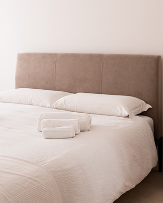 Un letto con la testiera in tessuto color talpa e gli asciugamani arrotolati sul copriletto bianco, in luce morbida.