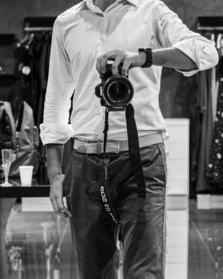 Un uomo in camicia bianca tiene con una mano la fotocamera appesa alla tracolla, in bianco e nero, davanti agli abiti appesi di un negozio.