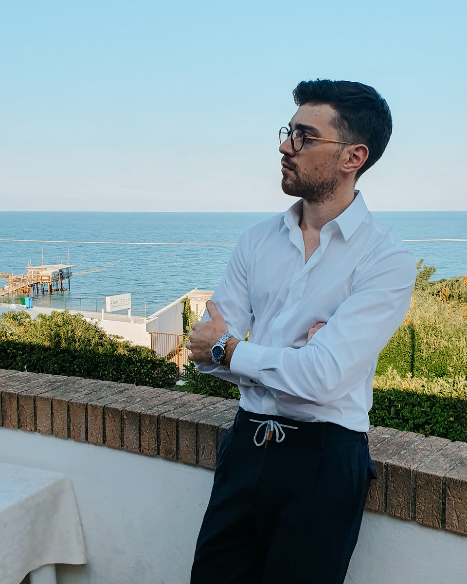 Ritratto di Piergiorgio Di Falco in camicia bianca e con gli occhiali, appoggiato a un muretto con le braccia conserte e lo sguardo verso sinistra; dietro di lui il mare e un trabocco.