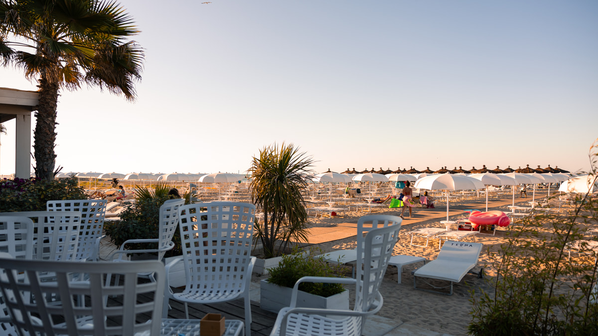 La terrazza di un locale sul mare al tramonto: sedie bianche vuote, una palma e, più in là, la spiaggia con gli ombrelloni.