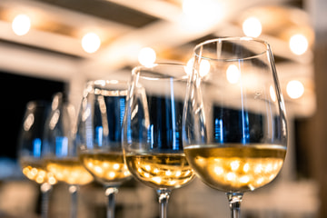Calici di vino bianco in fila e in diagonale, con le lampadine sfocate di un pergolato dietro.
