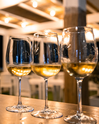 Tre calici di vino bianco in fila sul banco di un locale, con le luci calde di un pergolato sfocate dietro.
