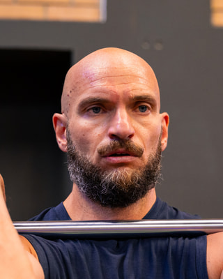 Un atleta con la barba e la testa rasata solleva un bilanciere all’altezza delle spalle, con lo sguardo intenso.