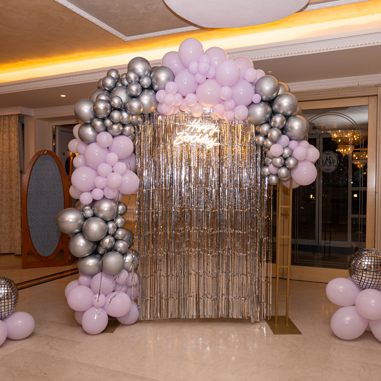 Un arco di palloncini lilla e argento, con due palle a specchio sul pavimento, davanti a una tenda di frange argentate, in una sala dalle luci calde.