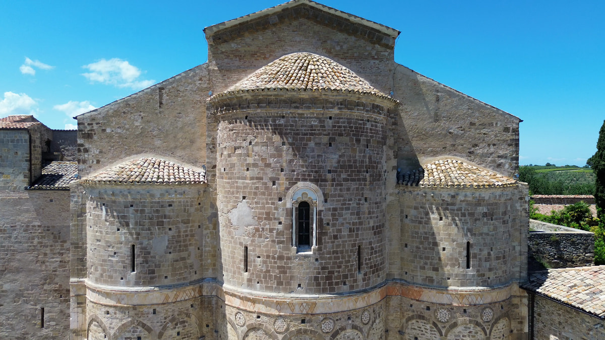 L’abside in pietra di una chiesa romanica vista dall’alto, con il tetto di coppi, i giardini intorno e il cielo azzurro.